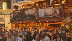 Menschenmenge auf einem belebten Weinfest vor einem zweistöckigen Holzstand mit beleuchteten Sonnenschirmen am Abend.