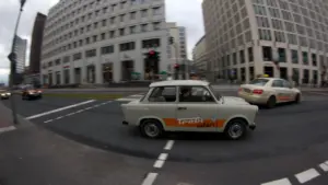Ein gelber Trabant fährt durch die moderne Berliner Innenstadt an Gebäuden vorbei.