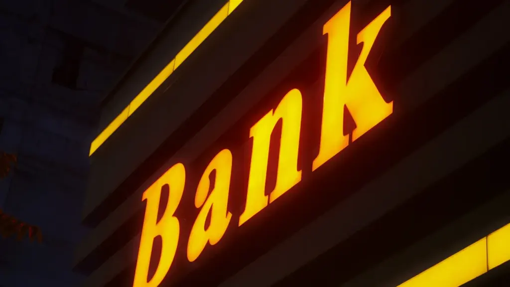 Ein beleuchtetes Firmenschild mit der Aufschrift Bank an einem Gebäude in der Dunkelheit.