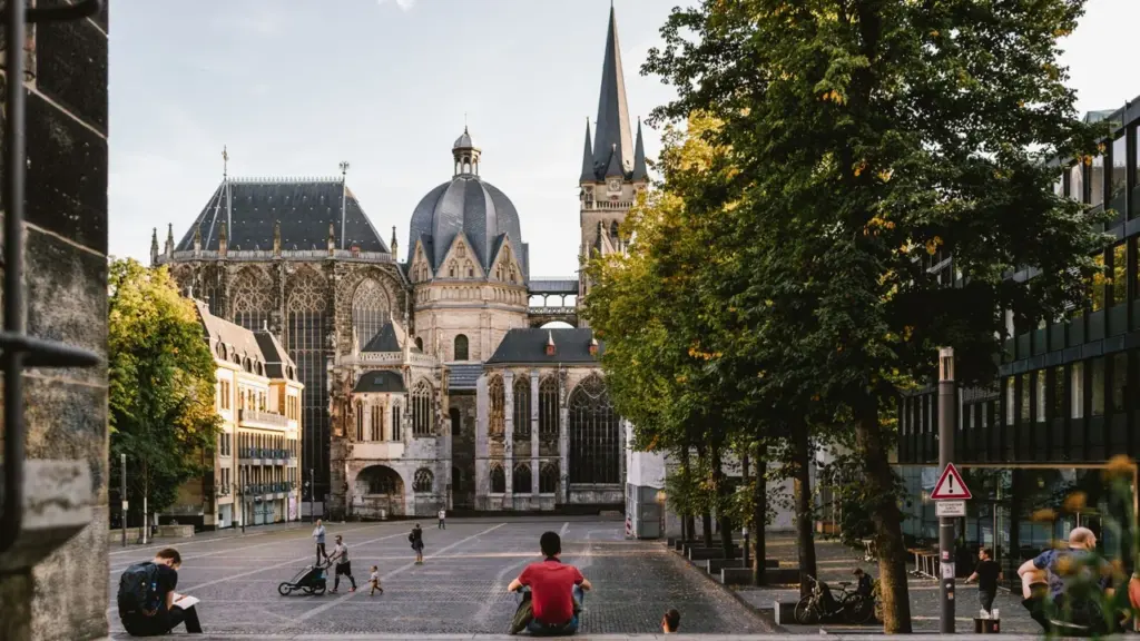 Blick auf den Aachener Dom bei sonnigem Wetter mit Menschen auf dem gepflasterten Vorplatz.