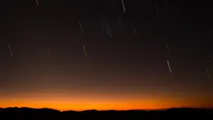 Sternschnuppen ziehen bei Sonnenuntergang als helle Lichtstreifen über die dunkle Silhouette eines Hügels am Nachthimmel.
