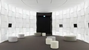 Ein futuristischer, heller Raum mit gekrümmten weißen Wänden und runden Sitzhockern auf dunklem Boden.