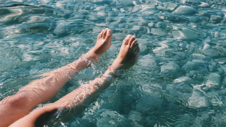 Beine einer Person, die in klarem, durchsichtigem Wasser über Kieselsteinen entspannt.