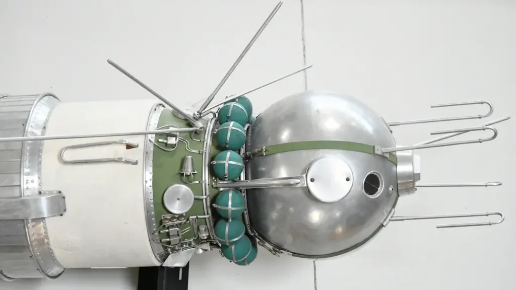 Ein historisches sowjetisches Vostok-Raumfahrzeug mit metallischer Kugelform und ausladenden Antennen vor grauem Hintergrund.